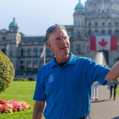 A man in a blue shirt gestures while standing outside near a historic building and a Canadian flag.