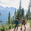Two hikers in a forested mountain area with cable cars overhead. Tall pine trees surround them under a clear sky.
