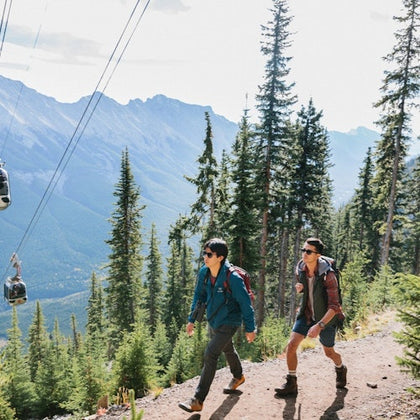 Two hikers in a forested mountain area with cable cars overhead. Tall pine trees surround them under a clear sky.