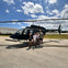 Four people standing in front of a parked helicopter on an airfield with buildings and a partly cloudy sky in the background.