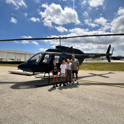 Four people standing in front of a parked helicopter on an airfield with buildings and a partly cloudy sky in the background.