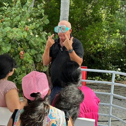 A man wearing diving goggles points to his eyes while speaking to a group of people, with lush greenery in the background.