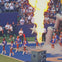 Cheerleaders in blue and white outfits perform on a football field with a large flame burst and a crowd in the background.