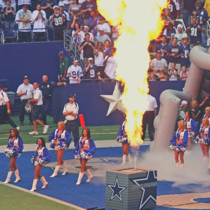 Cheerleaders in blue and white outfits perform on a football field with a large flame burst and a crowd in the background.