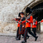 Guards in red uniforms and tall black hats march in formation near a stone archway.