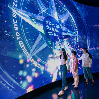 Three people interact with a large illuminated touchscreen displaying colorful graphics and Japanese text in a dark room.