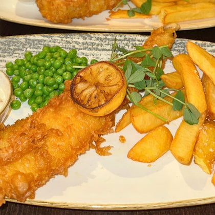 A plate with battered fish, lemon slice, peas, chips, and garnish on a light ceramic dish.