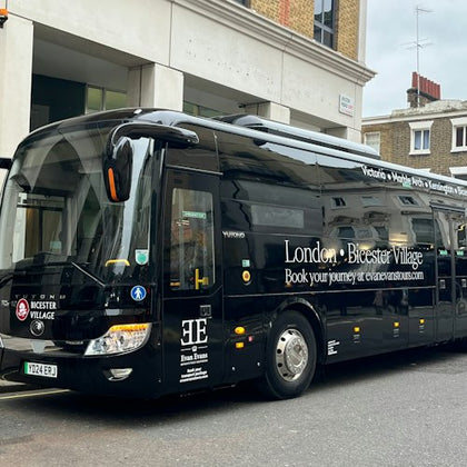 Black tour bus parked on a city street with "London - Bicester Village" and other text advertising a travel service on its side.