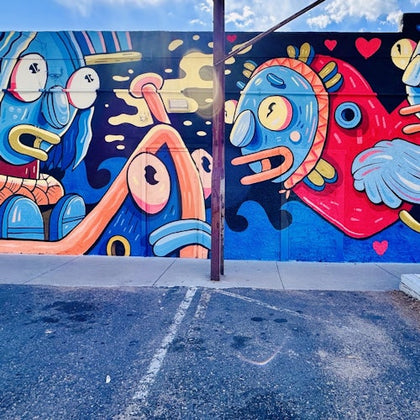 Colorful mural on a wall featuring abstract cartoon characters with vibrant hues and dynamic shapes, viewed from a parking lot.