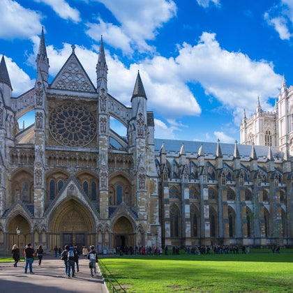 A view of a Gothic-style cathedral with ornate details, a rose window, and people walking in a grassy area under a blue sky.