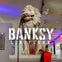 A lion statue on a pedestal displayed in an art gallery with the text 'BANKSY LIMITLESS' overlaid on the image.