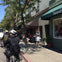 Cyclists riding along a street with shops and cafes, including one with a sign reading 'Sausalito,' and pedestrians nearby.