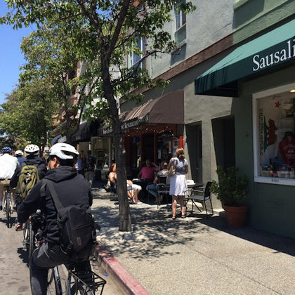 Cyclists riding along a street with shops and cafes, including one with a sign reading 'Sausalito,' and pedestrians nearby.