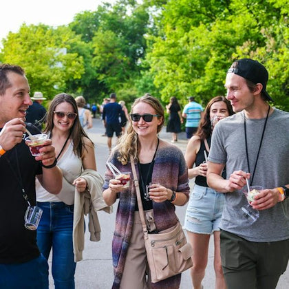 A group of people, some wearing sunglasses, are walking outdoors and enjoying food from cups on a sunny day. Trees are in the background.