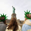 Three people wearing green Statue of Liberty crown hats face the Statue of Liberty, which stands on its pedestal.