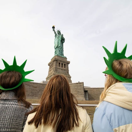 Three people wearing green Statue of Liberty crown hats face the Statue of Liberty, which stands on its pedestal.