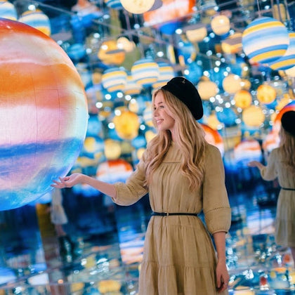 A woman in a beige dress and black beret poses in a reflective room filled with colorful globe-like decorations.