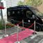 A black van is parked on a city street behind a red carpet setup with stanchions and red ropes.
