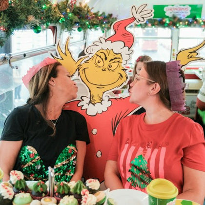 Two people seated at a festive table with a cardboard Grinch figure between them, surrounded by holiday decorations and treats.