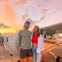 A smiling couple stands in front of a small airplane at sunset, with warm light and clouds in the sky.