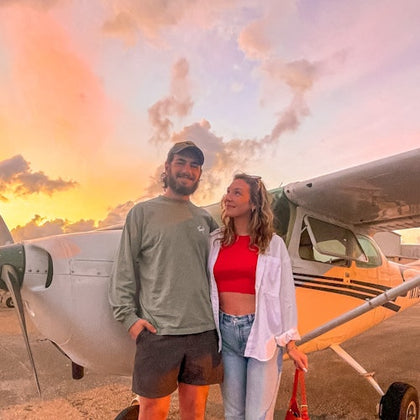 A smiling couple stands in front of a small airplane at sunset, with warm light and clouds in the sky.