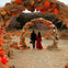 Two people walk hand in hand through an arch decorated with pumpkins and hay. Surrounding them are hay bales and more pumpkins.
