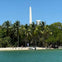 Beach with palm trees, a boat, people on the shore, and a tall white obelisk in the background under a clear blue sky.