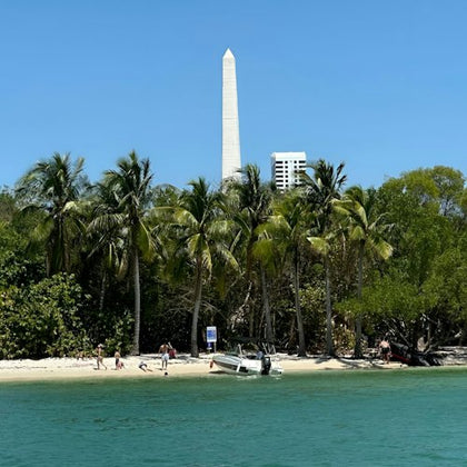 Beach with palm trees, a boat, people on the shore, and a tall white obelisk in the background under a clear blue sky.