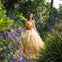 A woman in a flowing yellow gown stands in a lush garden surrounded by green foliage and purple flowers.