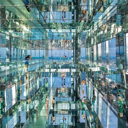 People walking in a mirrored room with glass walls, floors, and ceilings, reflecting the cityscape and themselves.