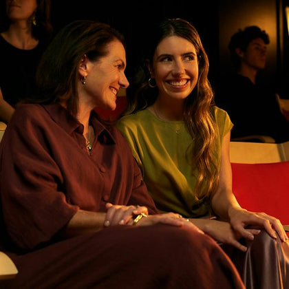 Two women smiling and sitting together in a warmly lit room, with others blurred in the background.