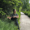 Person in a green shirt gesturing while standing beside tall grass on a path in a lush, forested area.