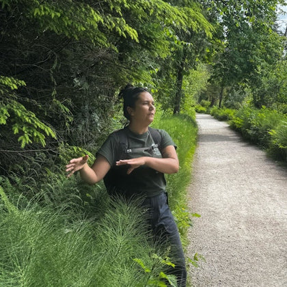Person in a green shirt gesturing while standing beside tall grass on a path in a lush, forested area.