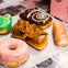 Five assorted donuts with various toppings, including pink glaze, bacon, chocolate swirl, green icing, and cream filling on a newspaper.