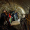 A group of people exploring an underground tunnel with a guide holding a flashlight, surrounded by aged and worn walls.