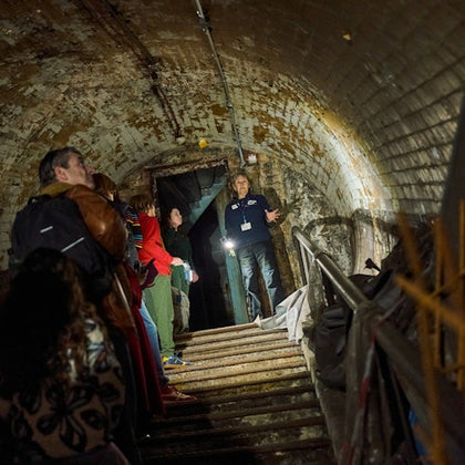 A group of people exploring an underground tunnel with a guide holding a flashlight, surrounded by aged and worn walls.