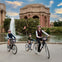 Three people with helmets ride bicycles near a fountain and a neoclassical building with columns and a dome.