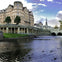 A historic building with columns by a river, ducks in the water, and a distant church spire under a partly cloudy sky.