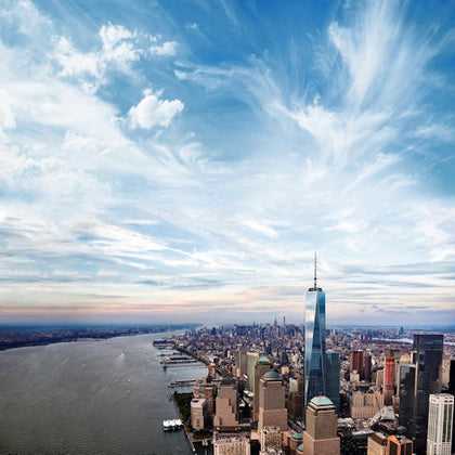 Aerial view of a cityscape featuring a prominent skyscraper, a river on the left, and a sky filled with wispy clouds.