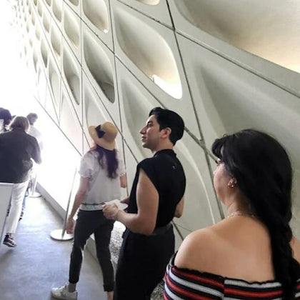 People walking in a brightly lit modern hallway with a patterned, geometric wall. One person wears a hat, and another has an off-shoulder top.