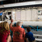 A group of people sitting and facing a presentation of a space module interior displayed in an exhibition setting.