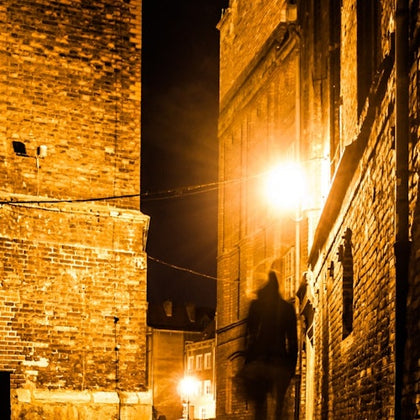 A person walking through a narrow alleyway at night, illuminated by orange streetlights and surrounded by brick buildings.