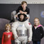 Four children posing with a space suit display; one child is inside the suit, another is climbing on top. Moon and Earth background.