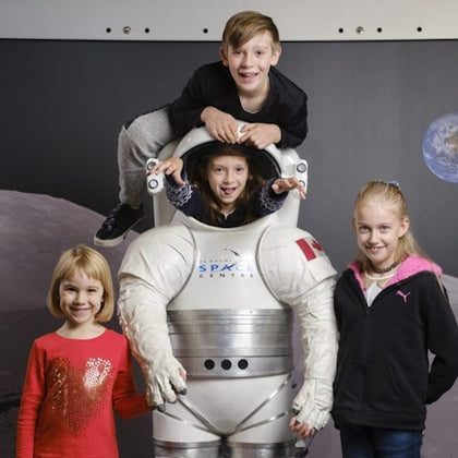 Four children posing with a space suit display; one child is inside the suit, another is climbing on top. Moon and Earth background.