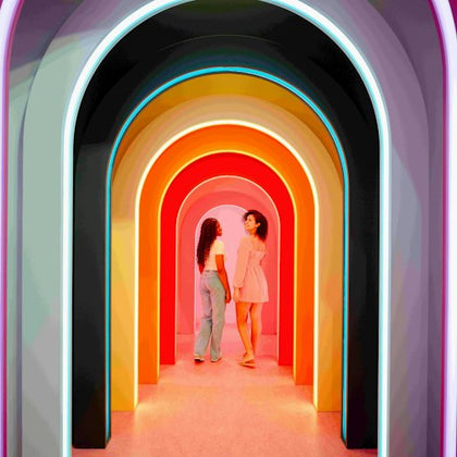 Two women standing under a series of colorful, illuminated archways, facing and smiling at each other.