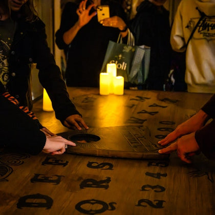 People using a Ouija board on a table with candles, letters, and numbers visible.
