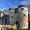 Historic stone castle with cylindrical towers under a clear blue sky with scattered clouds. People walking near the entrance.