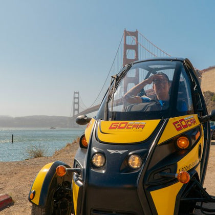 A person sits in a yellow and black GoCar with the Golden Gate Bridge visible in the background on a sunny day.