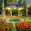 A colorful garden with red flowers, a small pond, and a yellow and blue arch structure surrounded by lush greenery.
