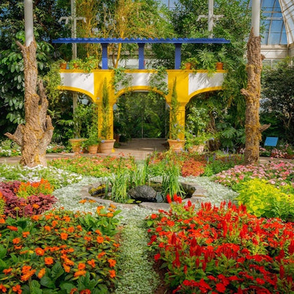 A colorful garden with red flowers, a small pond, and a yellow and blue arch structure surrounded by lush greenery.